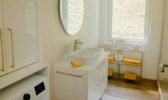 A modern bathroom with a washing machine and a sink. The design is bright and friendly, featuring a large mirror and a window view.