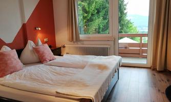 A cozy bedroom with a large double bed and pink pillows. Through the windows, one can see the green surroundings and the balcony.