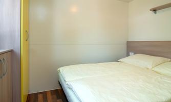 A simple bedroom with a double bed and a wardrobe. The walls are bright, and the floor is made of wood.