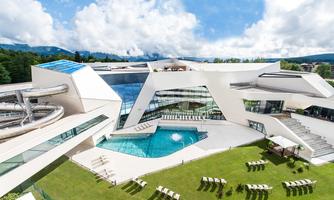 A modern building with interesting architecture and a pool. In the foreground, there is a green lawn area with loungers.