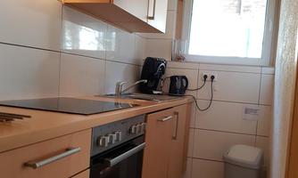 A modern kitchen with light wood furniture and white tiles. It is functionally furnished and has windows for daylight.