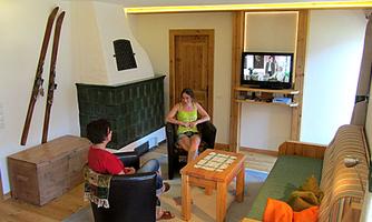 A cozy living room with a wood stove, television, and a dining table. Two people are sitting comfortably in armchairs and having a conversation.