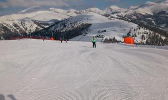 A snow-covered landscape with mountains in the background. Some skiers are enjoying the slope under a clear blue sky.