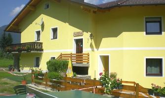 A light yellow house with a balcony and a colorful flower garden. The surroundings are green and inviting.