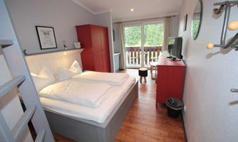 A cozy bedroom with a double bed and bright walls. The room has a balcony and wooden flooring, filled with natural light.