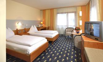 A cozy hotel room with two single beds and a television. There is a table with a floral arrangement and large windows that let in daylight.