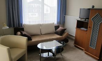 A cozy living room with a beige sofa, a glass coffee table, and a TV. The large window front lets in plenty of light.