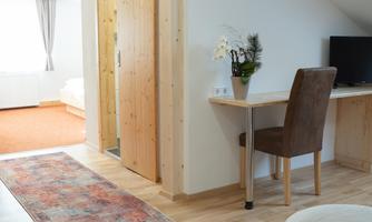 A cozy room with wooden floors and a desk. In the background, a bright bedroom with a carpet can be seen.