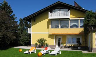 A yellow house with large windows and a balcony. In the foreground, there are sun loungers and garden balls on a green lawn.