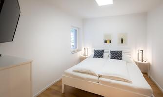 A modern bedroom with a double bed, white walls, and two nightstands. There is a window that lets in natural light, as well as a television on the wall.