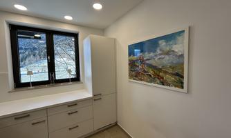A modern kitchen with white cabinets and a large window. A colorful landscape painting hangs on the wall.