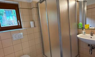 A bright bathroom with a shower cubicle and a sink. The walls are tiled, and there is a window that lets in daylight.