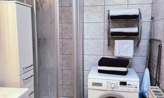 A modern bathroom with a shower and a washing machine. The walls are tiled with bright tiles and towels are arranged on a shelf.