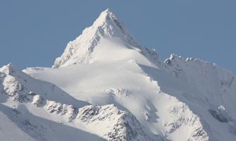 An impressive mountain peak with a snow-covered summit and clear blue sky. The surrounding mountains are also covered with snow.