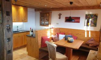 A cozy kitchen with wooden shelves and warm light.  
On a table with cushions sit some bottles of wine and a decorative bowl.