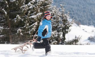 A child with a sled is happily running through the snow. The surroundings are wintry with green pine trees in the background.