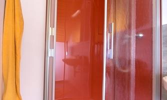 A modern shower cabin with a red wall and a white shower floor. A yellow towel hangs next to it.