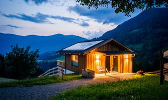 A cozy wooden house in the mountains, illuminated by warm light. The surroundings display a picturesque landscape with gentle hills and a clear evening sky.