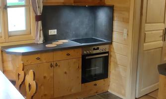 A cozy wooden kitchen with a modern stove and a sink. The floor is tiled with simple slabs and there is a large window with curtains.