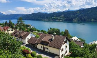 A picturesque view of a tranquil lake surrounded by mountains. In the foreground, cozy houses and plenty of green vegetation can be seen.
