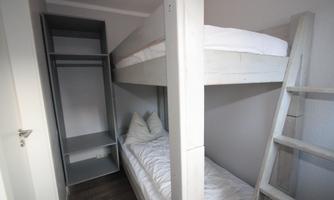 A simple room with a bunk bed and a single bed. There is a gray wardrobe and bright, neutral light.