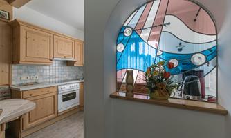A cozy kitchen with wooden cabinets and a window featuring colorful glass design. On the windowsill, there are flowers and a kettle.