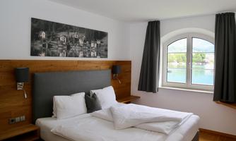 A modern bedroom with a large bed and elegant decoration. It has a window overlooking the water and is brightly lit.