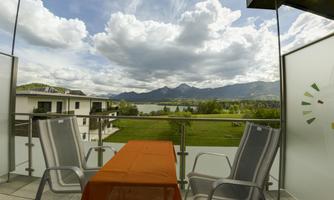 A beautiful balcony with two chairs and an orange table. In the background, mountains and a lake can be seen under a partly cloudy sky.