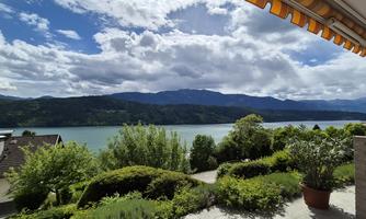 A beautiful view of a lake surrounded by mountains and wooded hills. The sky is partly cloudy, and it is a calm, peaceful day.