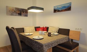 A cozy dining table with seating for four people. Pictures hang on the wall and a modern lamp provides the lighting.