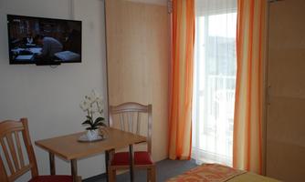 A cozy room with a table and chairs. A television is hanging on the wall and there is a balcony with curtains in orange and white.