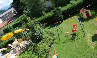 A green playground with a swing and playhouse. In the background, there are sunny terraces with chairs and umbrellas.