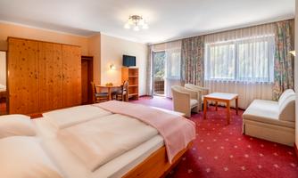 A bright hotel room with two beds, a table, and chairs. Large windows offer a view of nature and provide plenty of daylight.