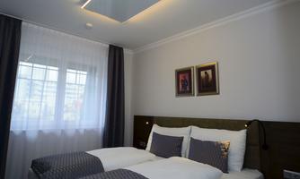 A modern bedroom with two beds, a stylish headboard, and soft colors. Large windows let in plenty of light, complemented by decorative pictures on the wall.