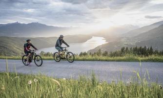 Two cyclists ride on a picturesque path overlooking a lake. In the background, mountains can be seen under a cloudy sky.