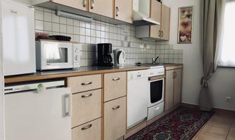 A modern kitchen with light wooden furniture and white appliances. There is a rug and windows that let in natural light.