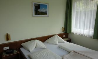 A simple room with two beds, white pillows, and a green curtain. A picture hangs on the wall and the window is brightly lit.