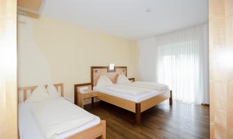 A bright bedroom with two beds and wooden flooring. Large windows provide plenty of daylight.