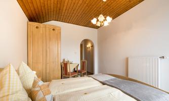 A cozy bedroom with two beds and wooden ceilings. In the background, a dining table with chairs can be seen.