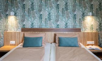 A modern bedroom with two single beds and stylish cushions. The wall is decorated with a tropical leaf pattern and there is soft lighting.