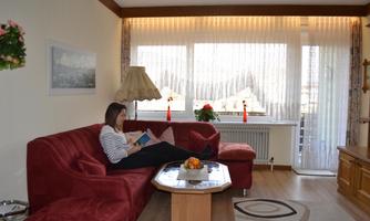 A cozy living room with a red sofa and a lamp. A person is sitting on the sofa reading, while daylight streams through the windows.