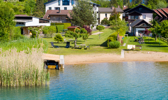 A peaceful lake landscape with well-kept gardens and popular houses. In the foreground, there is a sandy beach and boat docks.