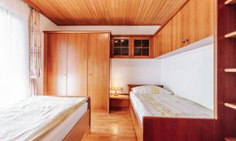 A cozy bedroom with two single beds and wooden paneling. There is a large wardrobe and plenty of daylight coming through the window.