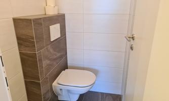 A modern bathroom with a white toilet and a small wall shelf. The walls are light tiled and the floor has a dark tiled surface.