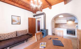 A cozy living room with a sofa and a dining table. The kitchen is modern and open-plan, featuring a wooden beam ceiling and warm lighting.