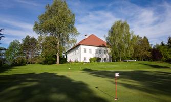 A well-maintained golf course with a picturesque 17th-century castle in the background. Tall trees surround the green landscape under a clear sky.