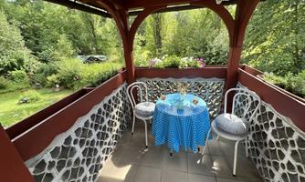 A cozy balcony with a blue table and two white chairs. In the background, green plants and flowers can be seen.