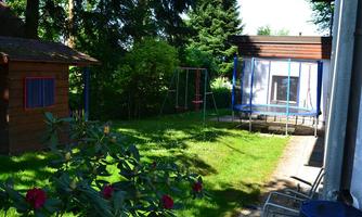A beautiful garden with a lawn, a trampoline, and a swing. In the background, there are trees and a wooden house.