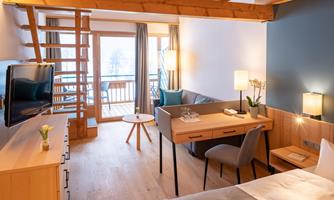 A cozy hotel room with wooden decor and a light color palette. There is a small desk, a chair, and a balcony with a view outside.