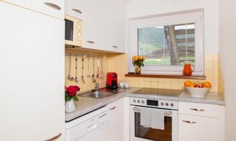 A bright and modern kitchen with white cabinets and a windowsill full of flowers and fruit. The equipment includes a stove, a microwave, and a coffee machine.